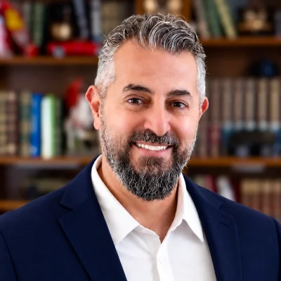 Smiling man with short gray hair and a beard wearing a navy suit and white shirt, standing in front of a blurred bookshelf filled with books and decorative items.