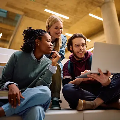 Three young adults sit on steps indoors, smiling and looking at a laptop together. They appear to be collaborating or studying in a modern, well-lit space.